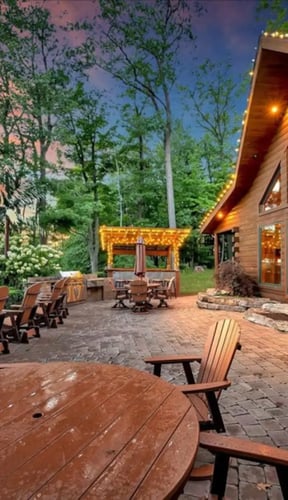 Lakeside log cabin at dusk with string lights, stone patio, and Adirondack chairs by the water